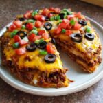 A close-up of a layered mexican pizza, topped with melted cheddar cheese, black olives, diced tomatoes, and green onions.