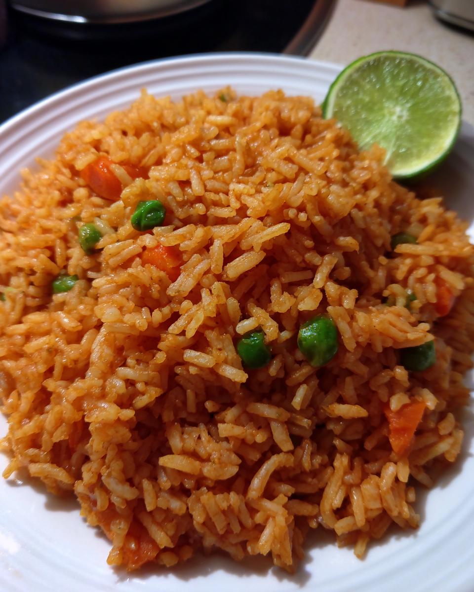 A close-up of fluffy, orange-colored Mexican rice recipe served on a white plate, garnished with peas, carrots, and a lime wedge.
