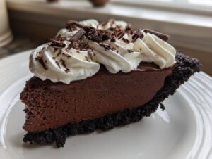 A decadent slice of French silk pie featuring a dark chocolate mousse filling and a chocolate cookie crust, topped with whipped cream and shavings.