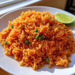 A close-up of a mound of vibrant orange Mexican rice recipe, mixed with green peas, served on a white plate with a lime wedge.